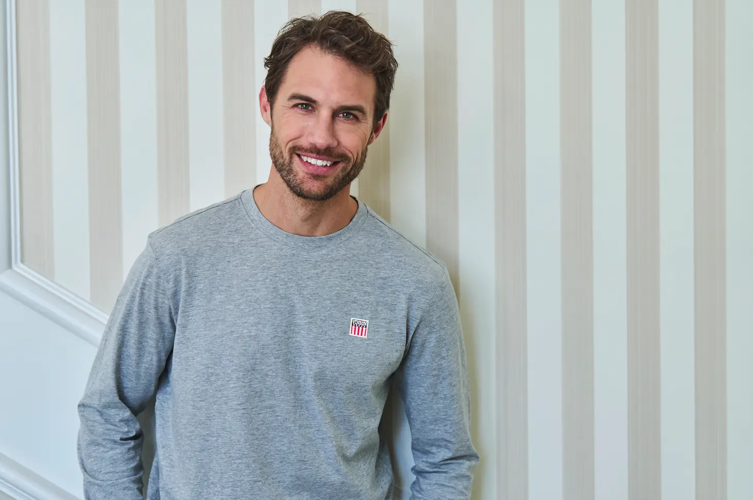 Hombre con sudadera gris de cuello redondo junto a una pared de rayas blancas - Dress code casual hombre VINSON